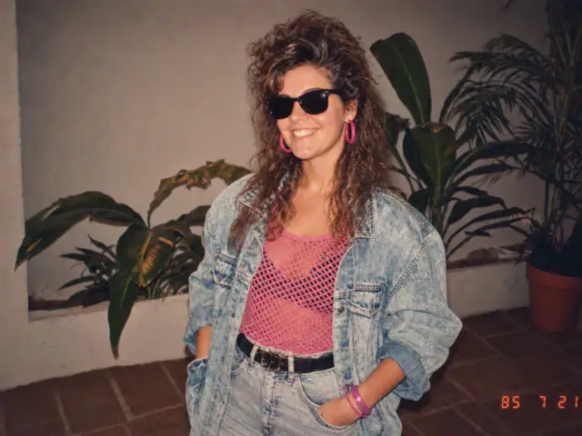 A smiling woman in black sunglasses, bright pink hoop earrings, a pink mesh top, and an oversized denim jacket poses in front of indoor plants. A red-orange date stamp reads “85 7 21” in the lower-right corner.